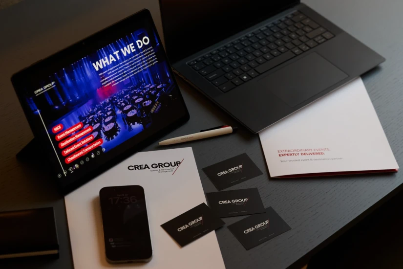 Mesa de trabajo de CREA Group con ordenador portátil, tablet mostrando los servicios de gestión de eventos MICE, teléfono móvil, tarjetas de visita y papelería corporativa.