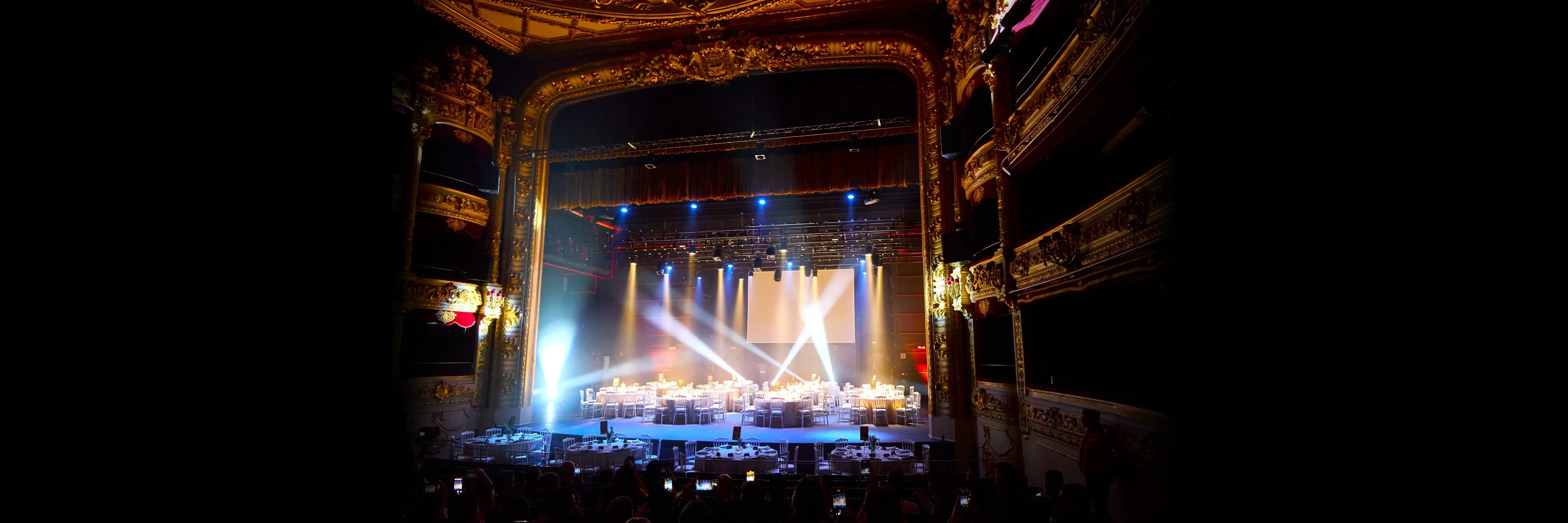 Vista panoràmica d'un sopar de gala muntat a l'escenari d'un teatre històric amb il·luminació dramàtica.
