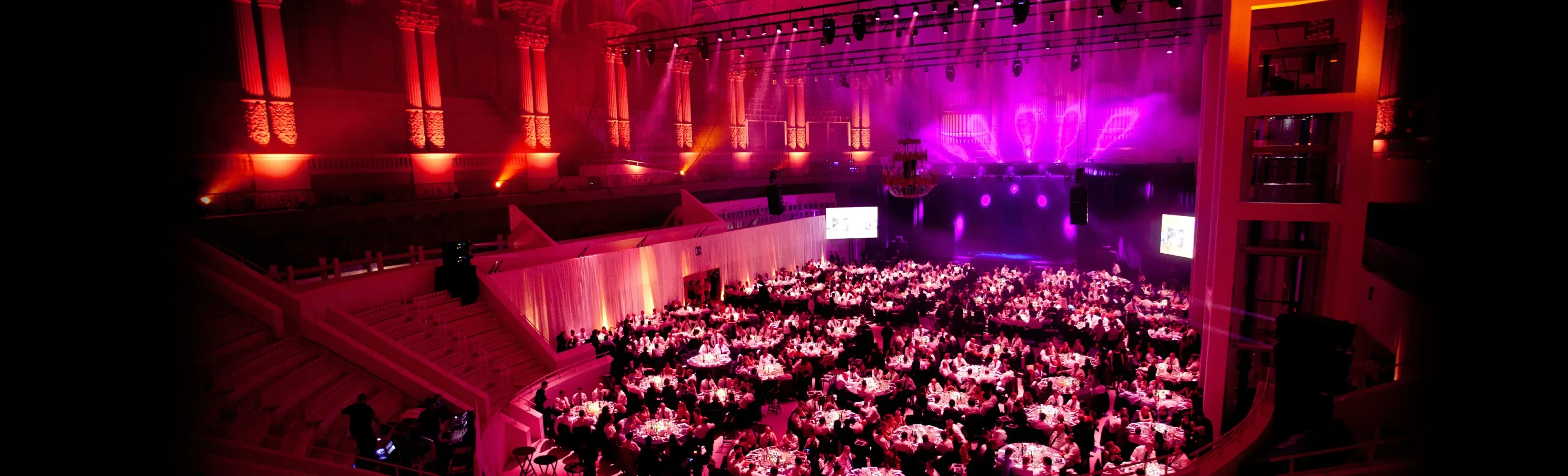 Vista panorámica de una gran cena de gala corporativa con una iluminación de escenario vibrante en tonos rosa y púrpura.
