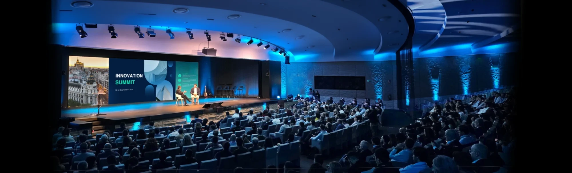 Gran auditorio de convenciones corporativas con escenario de Innovation Summit e iluminación azul.