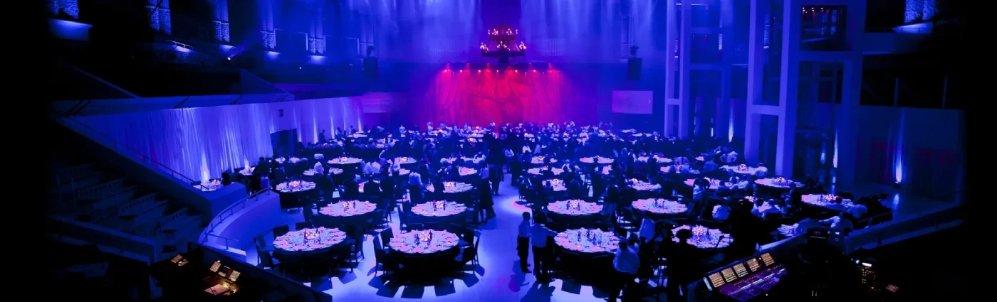 Salle de bal élégante aménagée pour banquet avec éclairage bleu et violet, tables rondes remplies d'invités, grande scène à fond rouge et hauts plafonds pour un gala.