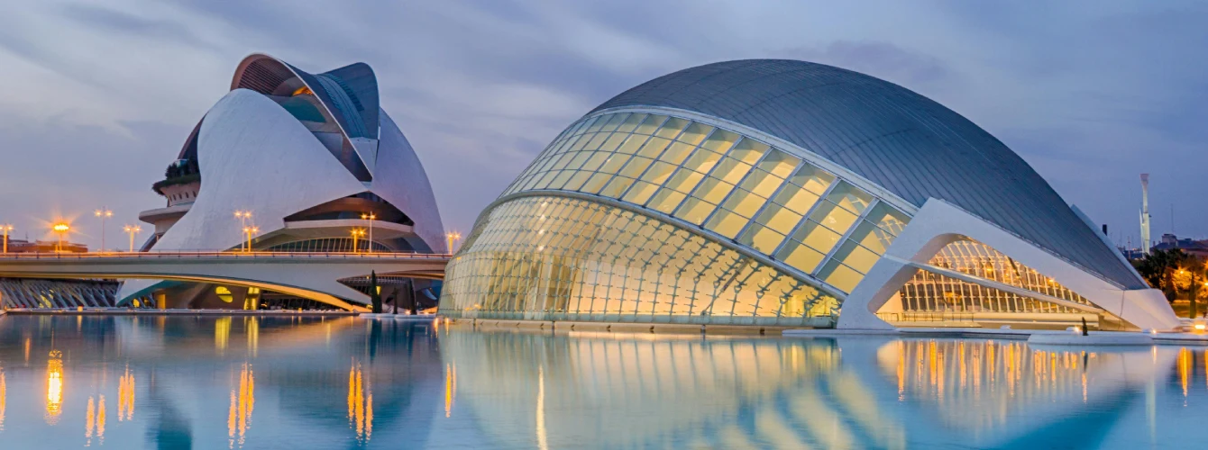 Night view of the illuminated Hemisfèric, shaped like an eye, and the Palau de les Arts Reina Sofía at Valencia’s City of Arts and Sciences, reflected in still water beneath a twilight sky with warm lights