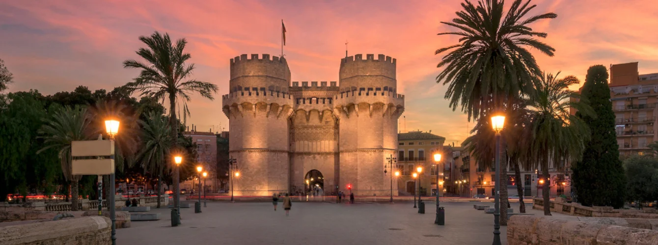 Sunset view of the Torres de Serranos, the iconic medieval city gates in Valencia, Spain, illuminated warmly against a vibrant pink and orange sky. Tall palm trees frame the scene on both sides, with classic street lamps glowing in the foreground and a few pedestrians walking across the spacious plaza in front of the historic twin-towered stone fortress. The ancient architecture features battlements, cylindrical towers, and intricate details, capturing the romantic atmosphere of Valencia at dusk.