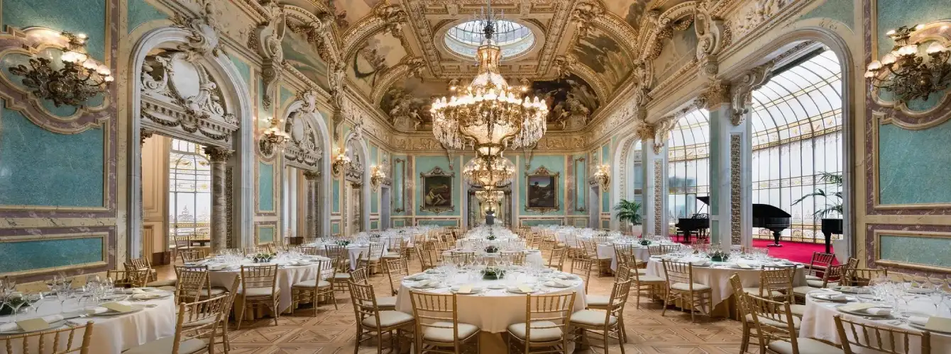 Opulent ballroom at night with tall arched stained-glass windows, warm golden ambient lighting, intricate wall decorations, and multiple round tables elegantly set with white tablecloths, glassware, silverware, and gold chairs, creating a luxurious atmosphere for a formal evening banquet or celebration.