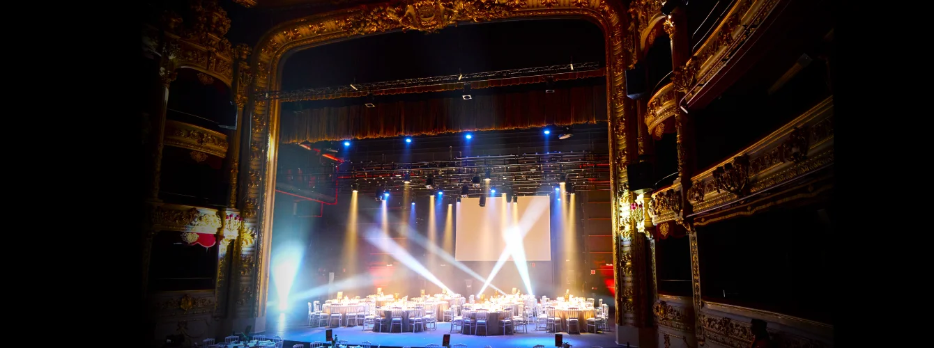 View from the stage of a historic theater featuring elaborate golden moldings, ornate balconies, and proscenium arch, with the auditorium floor transformed into a luxurious banquet hall filled with round tables illuminated by dramatic spotlights and surrounded by chairs for an exclusive sophisticated event.