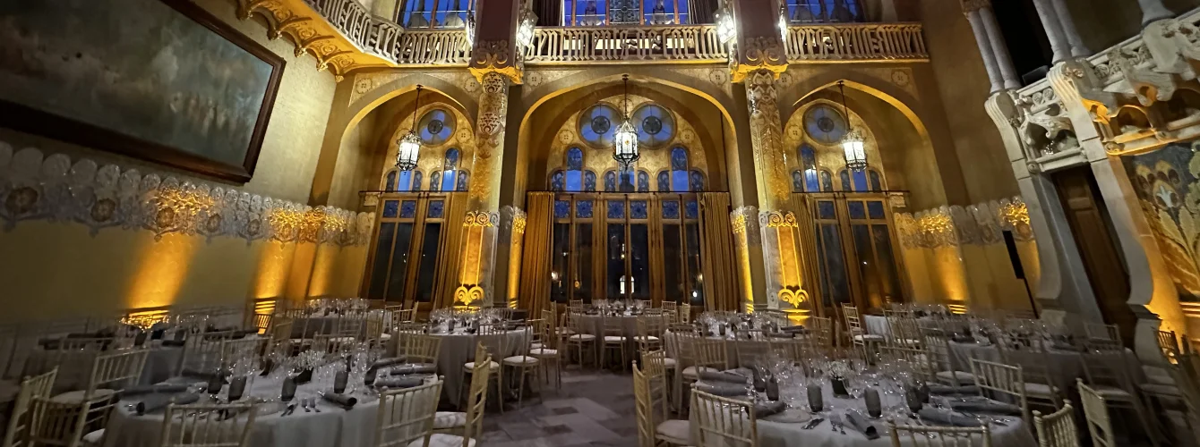 Historic ornate ballroom with high frescoed ceilings, massive crystal chandeliers, turquoise and gold wall panels, geometric parquet flooring, and round banquet tables dressed in white linens with gold chairs and floral centerpieces, prepared for an elegant gala dinner with grand pianos in the background.