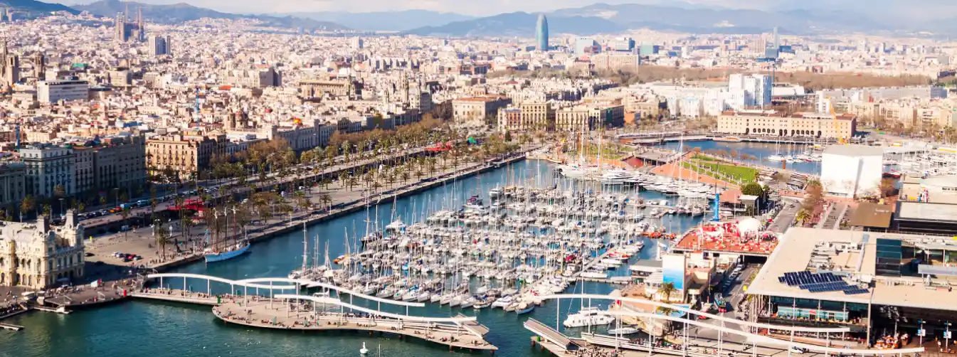 Scenic daytime view of Port Vell harbor in Barcelona, capturing luxury yachts docked at the marina with the calm Mediterranean sea and the city's coastal skyline in the background, representing a prime nautical tourism destination