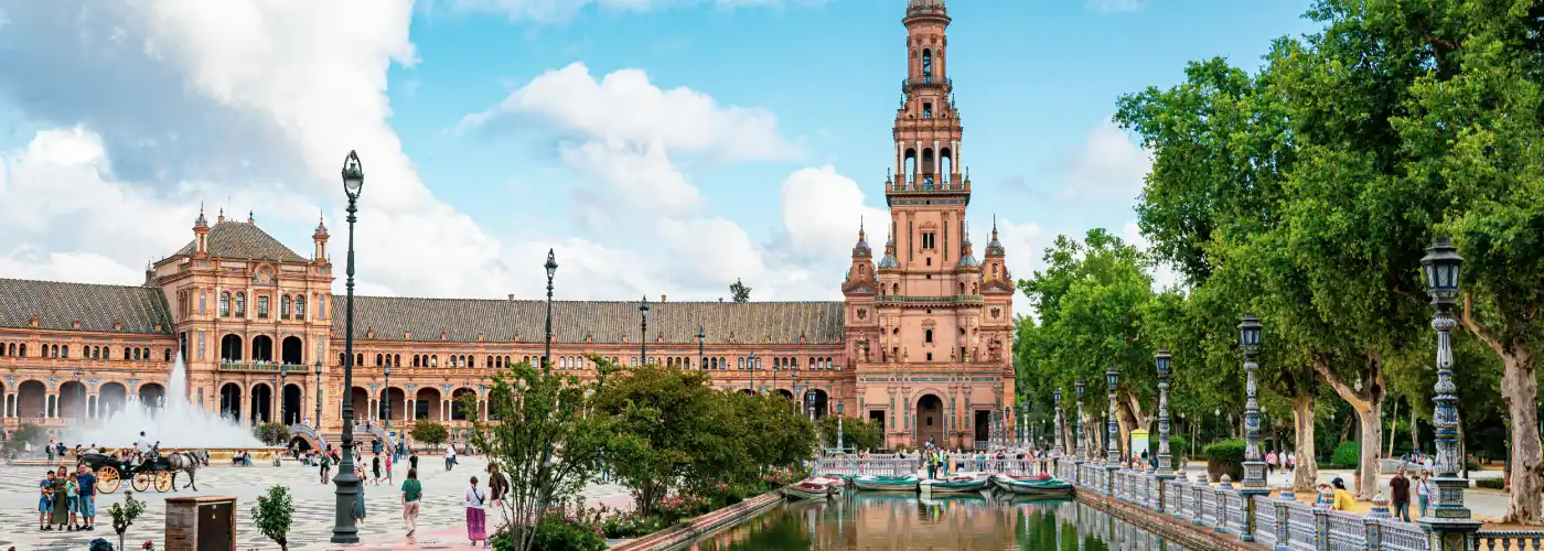 Breathtaking daytime view of the Plaza de España in Seville, showcasing the grand Renaissance Revival brick building and its tall tower reflecting in the canal, framed by ornate ceramic bridges and lush green trees under a dynamic blue sky, representing a top Spanish tourist destination