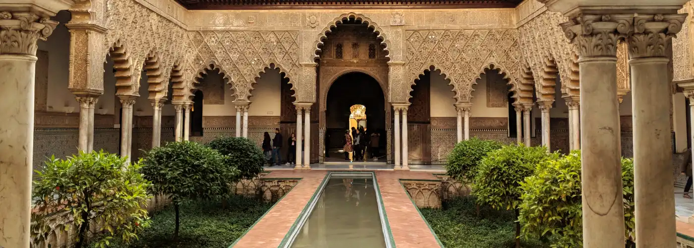 Exquisite Mudejar architecture of the Patio de las Doncellas in the Real Alcázar of Seville, featuring the central rectangular reflecting pool, intricate scalloped arches with detailed plasterwork, and small orange trees in a symmetrical sunken garden under a clear sky.