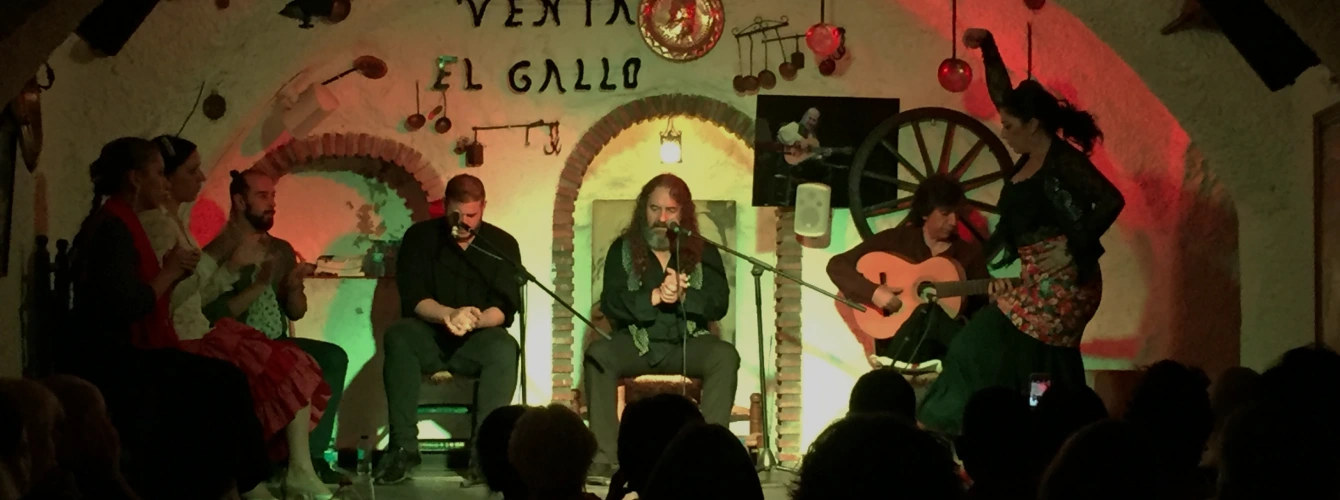 Passionate flamenco performance at Venta El Gallo in the Sacromonte caves of Granada, showing a male singer and guitarist alongside a female dancer in a traditional dress, performing on a small stage with rustic cave walls and dim red lighting