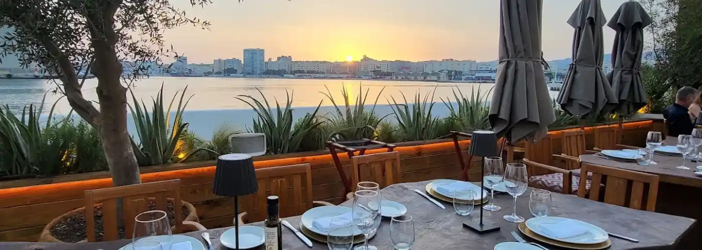 Elegant outdoor waterfront dining terrace at sunset with beautifully set wooden tables, wine glasses, and warm ambient lighting overlooking a serene bay and city skyline, perfect for romantic dinners