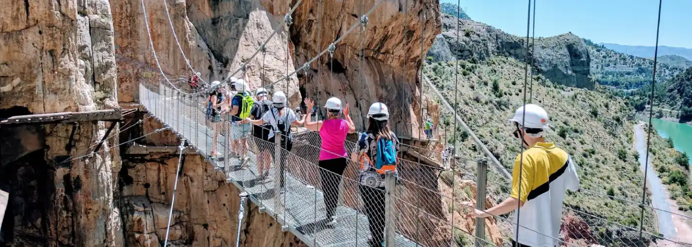 Group of tourists wearing white safety helmets walking single file along the thrilling Caminito del Rey suspension bridge attached to steep rocky cliffs with stunning mountain and reservoir views in Andalusia, Spain