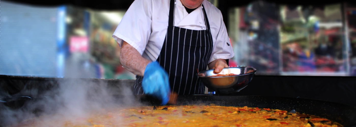 Professional chef in white uniform and blue gloves stirring a large traditional Spanish seafood paella in a giant pan with rising steam at an outdoor market or restaurant in Spain