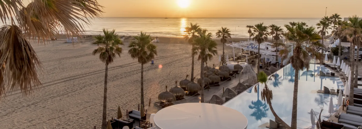 Luxurious beachfront club at sunset featuring an infinity pool with white sun loungers and thatched umbrellas, surrounded by tall palm trees overlooking a golden sandy beach and calm Mediterranean Sea in Spain