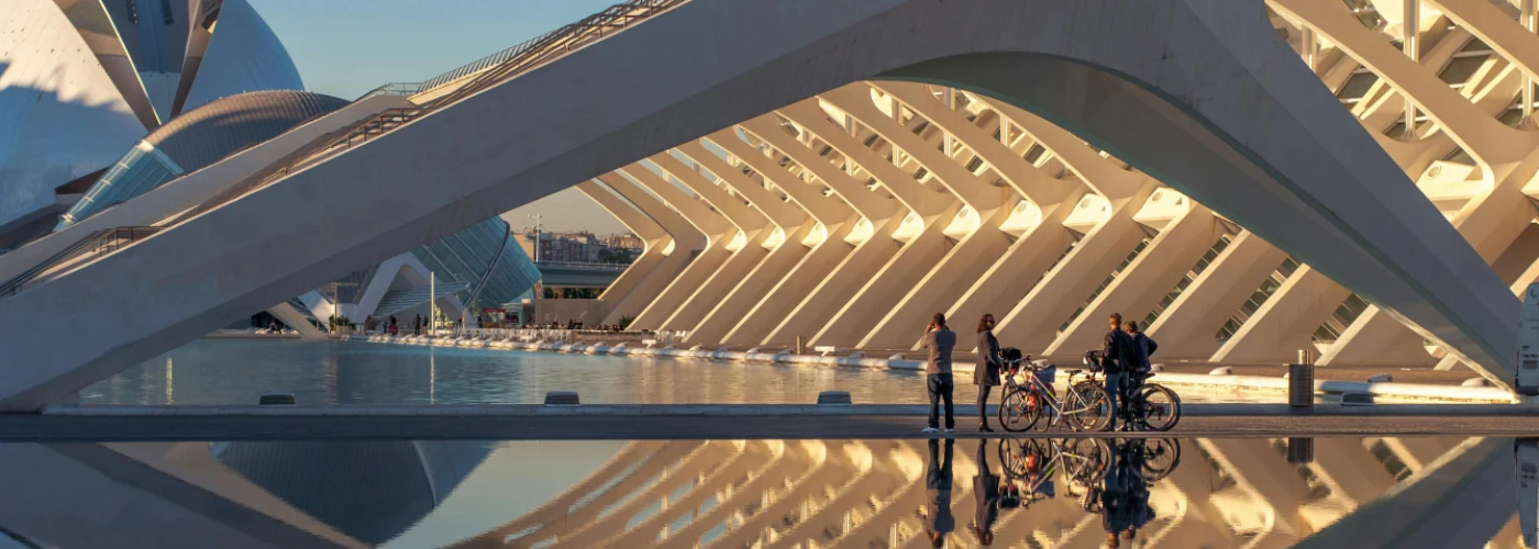 Modern architectural masterpiece of Valencia's City of Arts and Sciences with curved white structures and geometric patterns beautifully reflected in calm water, cyclists enjoying the scenic waterfront promenade at golden hour