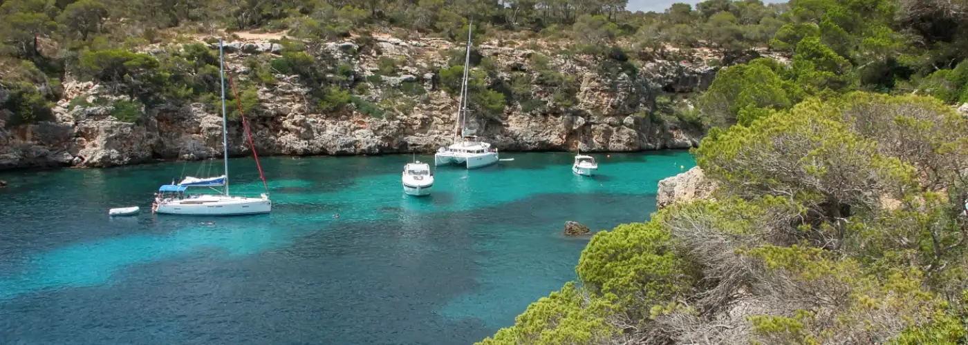 Idyllic secluded cove in Mallorca with crystal-clear turquoise waters, several anchored white sailboats and catamarans surrounded by dramatic rocky cliffs and dense Mediterranean pine forests
