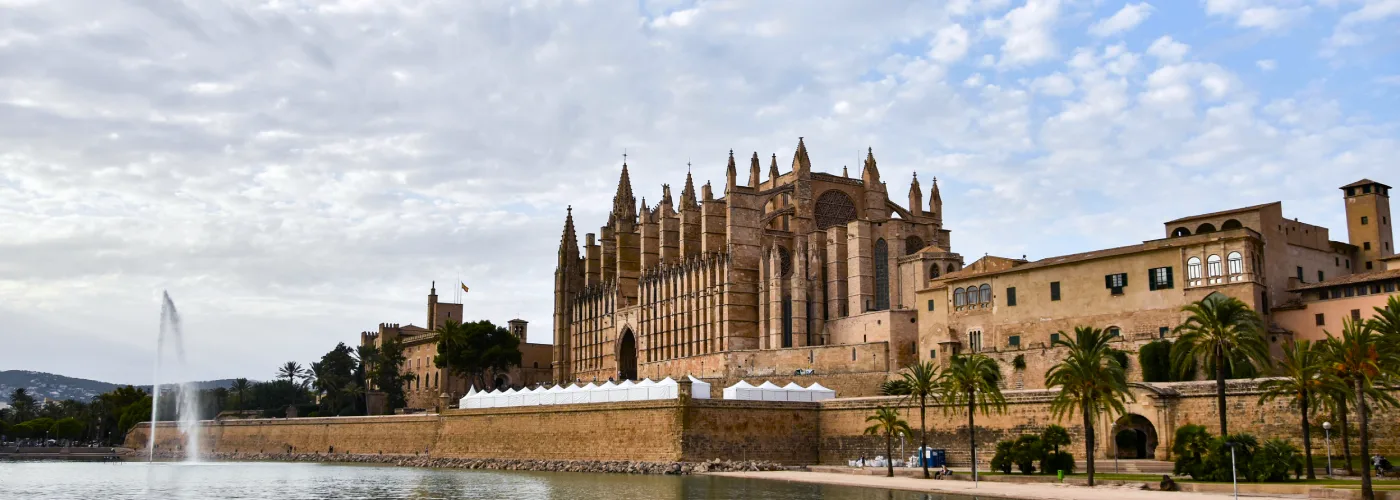 Majestic Gothic Cathedral of Santa Maria of Palma (La Seu) in Mallorca with intricate sandstone façade and towering spires reflected in calm waters of Parc de la Mar under a partly cloudy sky, surrounded by palm trees