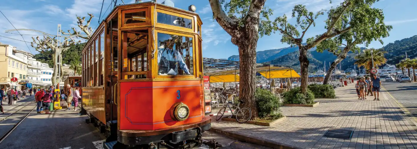 Historic orange wooden tram number 3 running along the scenic waterfront promenade in Port de Sóller, Mallorca, with passengers enjoying views of the marina, mountains and outdoor cafés on a sunny day