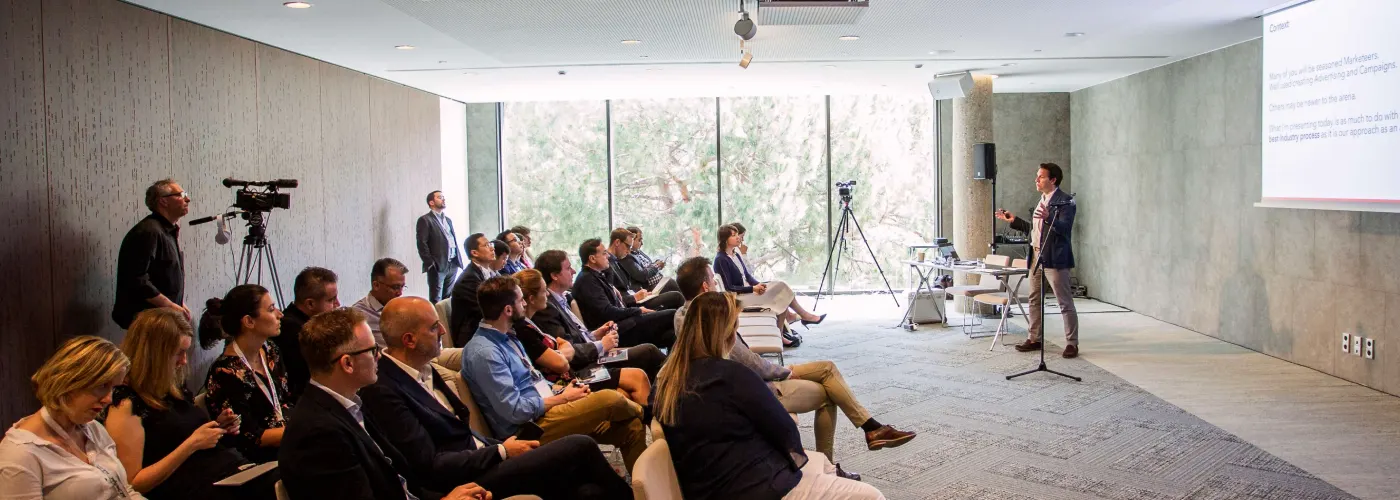 A presenter in a blazer gestures while speaking to an audience in a bright, minimalist seminar room. A large projection screen displays a "Context" slide, and large windows in the background show green trees. A videographer is filming the session from the side.