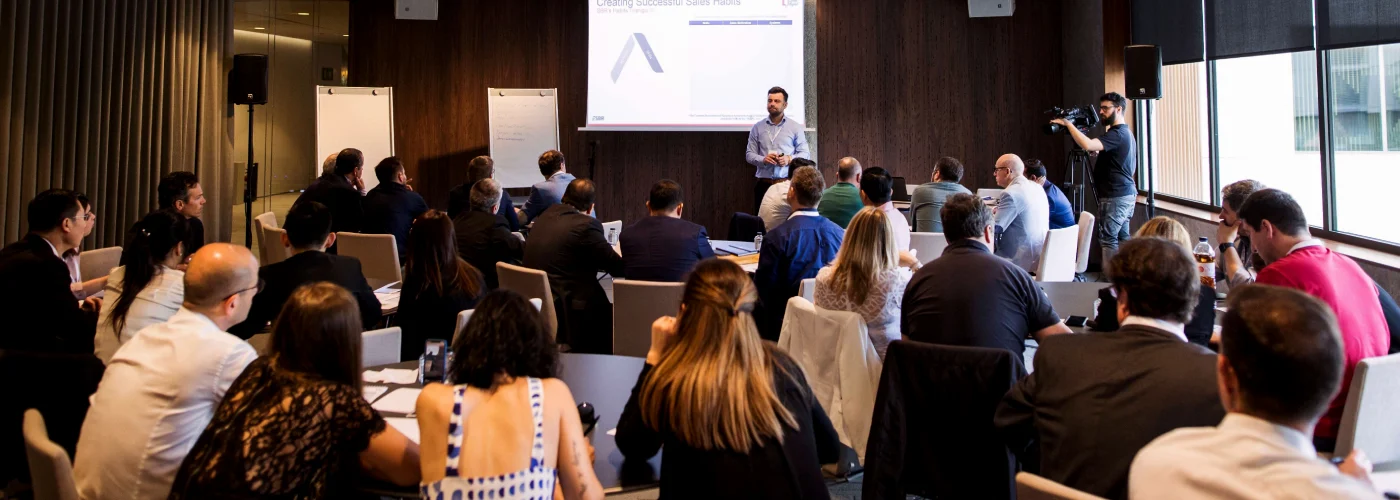 A male speaker stands in front of a projector screen titled "Creating Successful Sales Habits" during a corporate workshop. A diverse group of professionals sits at round tables, listening and taking notes in a modern conference room with dark wood paneling.