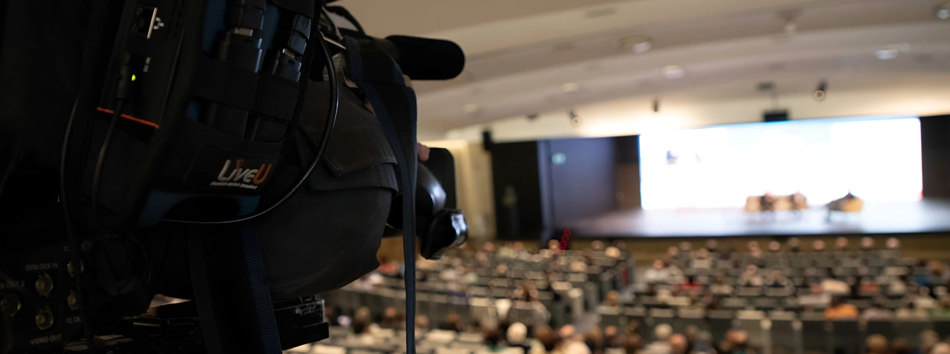 Close-up of a professional video camera with a LiveU unit broadcasting a conference in a large auditorium.