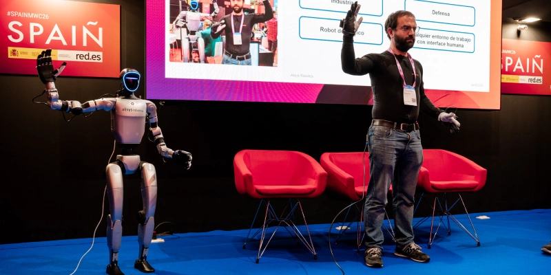 Un présentateur sur une scène au MWC Barcelone faisant une démonstration de suivi de sujet avec un robot humanoïde d'Alisys Robotics, devant un écran affichant des applications techniques.