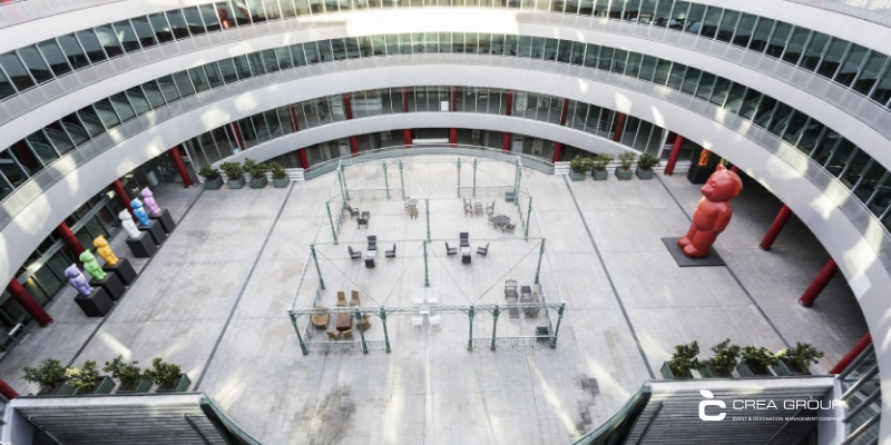 ifema-madrid-demo-bear-statues-indoor-courtyard Aerial view of the circular indoor atrium at IFEMA Madrid, featuring permanent installations of giant colorful bear sculptures by artist dEmo in vibrant colors (red, yellow, blue, green, and pink) arranged around the space. In the center, a seating area with tables, chairs, and green metal structures under a pergola, surrounded by potted plants and red columns. The modern design stands out with its multi-level glass galleries and abundant natural light from the ceiling. Photo taken from an upper level, showcasing the spaciousness and contemporary architecture of the Madrid exhibition center.